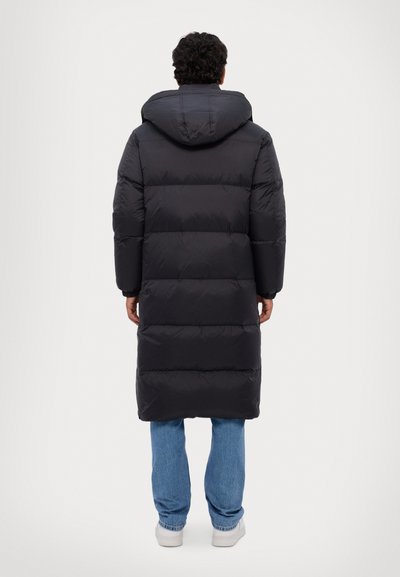 Lange zwarte pufferjas met een capuchon, voorzien van horizontale vulling, een ontspannen pasvorm en reikt tot halverwege de kuit, gecombineerd met lichtblauwe jeans.