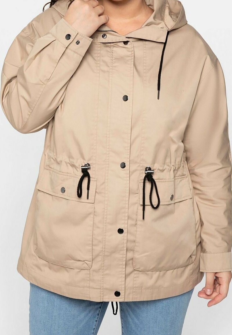 Beige Kapuzenjacke aus leichtem Material, mit schwarzen Kordeln, Druckknöpfen und zwei vorderen Taschen mit gesicherten Klappen.