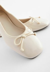 Ballerines ivoire avec un bout arrondi, en matière synthétique texturée et un nœud décoratif. Présentent une doublure intérieure lisse.
