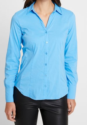 Femme portant une chemise bleu clair ajustée avec boutons, manches longues, et un pantalon en cuir noir sur un fond uni.