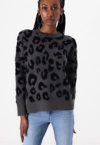 Maglione grigio con stampa leopardata nera, tessuto testurizzato, polsini e orlo a coste, scollatura rotonda e una vestibilità rilassata. Indossato con jeans blu.
