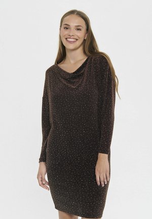 Šypsena moteris su ilgais plaukais, vilkinti blizgią tamsiai rudą ilgo rankovėmis suknelę su drapiruotu dekolte, stovi prieš paprastą baltą foną.