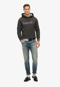 Sudadera gris oscuro con el logotipo de 'Superdry', bolsillo delantero, llevada sobre una camisa de rayas azul claro. Jeans azul claro, zapatos negros y reloj como accesorio.