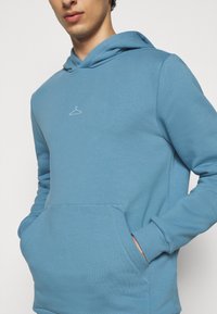 Jeune homme portant un sweat à capuche bleu avec une petite icône de cintre blanc sur la poitrine, les mains dans la poche avant, fond neutre.