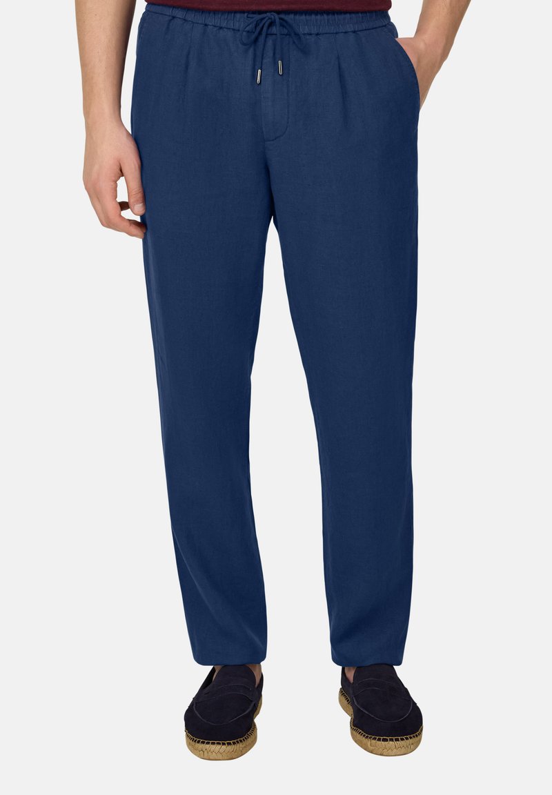 Homme portant un pantalon décontracté bleu marine avec cordon de serrage et des chaussures sombres sans lacets, debout avec une main dans la poche et un fond neutre.