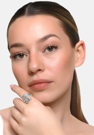 Młoda kobieta z gładką skórą i związanymi włosami, nosząca duży abstrakcyjny srebrny pierścień na palcu wskazującym blisko ust.