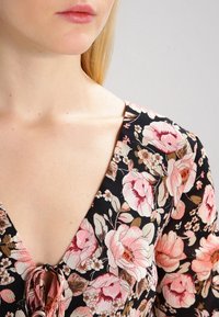 Černé květinové šaty s velkými růžovými a krémovými květy, s hlubokým výstřihem do V a šňůrkovým detailem u krku, vyrobené z lehkého materiálu.