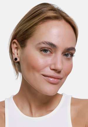Orecchini a perno neri su una persona dalla pelle chiara con capelli biondi corti, che indossa un top senza maniche bianco, sorridendo con un trucco naturale.
