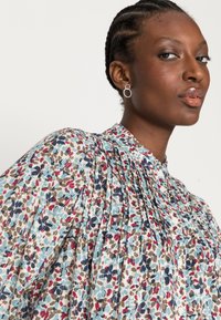 Blusa floral com pregas, apresentando um fundo azul claro e padrões multicoloridos em vermelho, castanho e azul. Gola alta com botões.