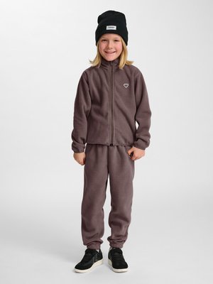 Uśmiechnięte dziecko ubrane w brązową polarową kurtkę i spodnie, czarną czapkę i czarne trampki, stojące na jednolitym białym tle.