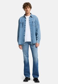 Chemise en denim bleu clair avec deux poches poitrine, associée à un jean bleu fuselé et des chaussures noires. T-shirt blanc porté en dessous.
