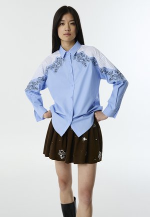 Camicia azzurra chiara a maniche lunghe con ricamo nero floreale e spalle a strisce, abbinata a una gonna plissettata marrone scuro con decorazioni.