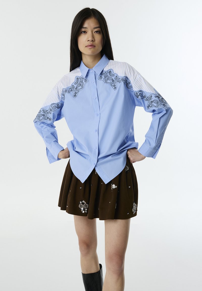 Camicia azzurra chiara a maniche lunghe con ricamo nero floreale e spalle a strisce, abbinata a una gonna plissettata marrone scuro con decorazioni.