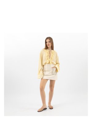 Młoda kobieta stojąca, ubrana w bluzkę w jasnym żółtym kolorze z falbankami, kremową mini spódnicę z kieszeniami wykończonymi koronką oraz beżowe, plecione sandały na płaskim obcasie na białym tle.