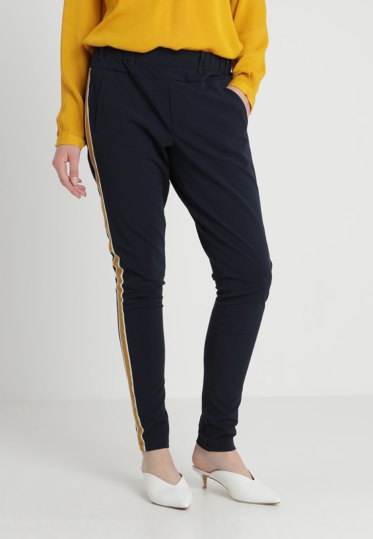 Pantalons slim-fit bleu marine en tissu extensible, avec des rayures dorées sur les côtés et des poches avant. Portés avec des mules à talons blancs.