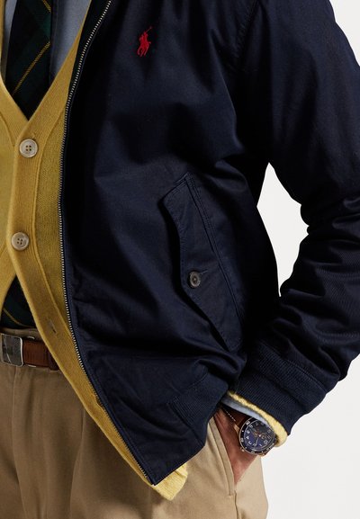 Marineblaue Zip-Jacke mit rotem Logo, senfgelbe Strickjacke darunter, beiges Paar Hosen und eine blaue Uhr mit einem Silberarmband.