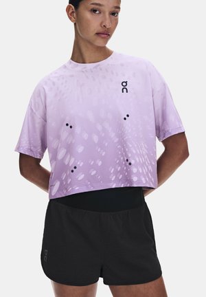 Donna che indossa una maglietta sportiva lavanda larga con puntini decorati e pantaloncini da corsa neri, in piedi con le mani dietro la schiena.
