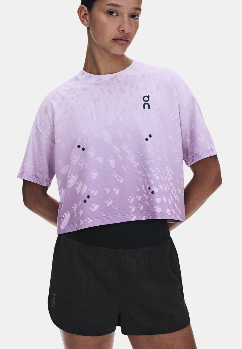 Femme portant un t-shirt de sport ample lavande avec des points à motifs et un short de course noir, debout avec les mains dans le dos.