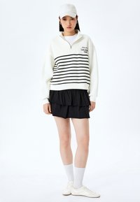 Witte zip-up sweatshirt met zwarte horizontale strepen, gelaagde zwarte gerimpelde rok, witte sokken, witte sneakers en een bijpassende pet.