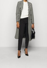 Abrigo largo de cuadros en blanco y negro con cuello, que se lleva sobre un suéter de punto blanco y pantalones negros, combinado con tacones negros y un bolso.