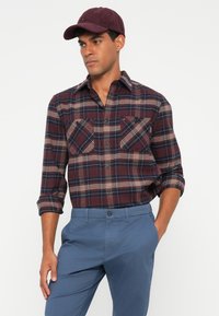 Camicia di flanella a quadri bordeaux con due tasche sul petto, maniche lunghe arrotolate, abbinata a pantaloni blu chiaro e un cappello bordeaux.