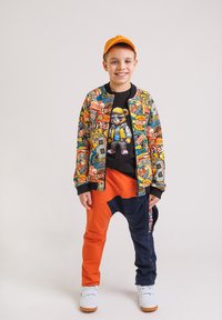 Uśmiechnięty chłopiec noszący pomarańczową czapkę, kolorową kurtkę z łatami sportowymi, czarną koszulkę z grafiką niedźwiedzia, dwu-tonowe pomarańczowo-granatowe spodnie oraz białe trampki.