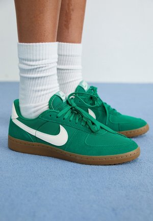 Zapatillas de gamuza verde y textil con el logo Nike swoosh blanco, suela de goma texturizada y cordones planos, usadas con calcetines blancos acanalados.