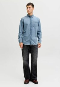 Hellblaues Jeanshemd mit Knöpfen und einer Brusttasche, kombiniert mit dunkelblauen Jeans mit geradem Bein und braunen Schuhen.