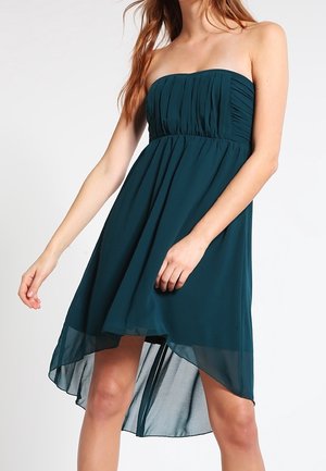 Robe bustier bleu sarcelle foncé à taille haute-basse avec corsage plissé portée par une femme, jupe fluide plus courte devant et plus longue derrière.