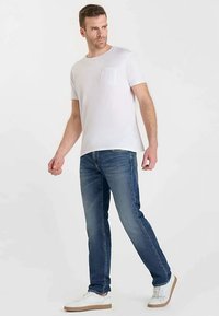 T-shirt blanc à manches courtes avec une poche sur la poitrine, associé à un jean en denim bleu. Les chaussures sont blanches avec une semelle beige, illustrant un style décontracté.