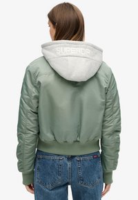 Grøn bomberjakke med grå hætte, ribbede manchetter og kant. Stoffet er glat med en let blank finish, og hætten har broderet tekst.