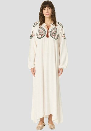 Femme debout portant une longue robe blanche avec une broderie florale sur les épaules et la poitrine, associée à des ballerines tissées beiges.