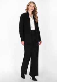 Schwarze offen front cardigan aus weichem Stoff, kombiniert mit einer weißen Satinbluse und schwarzen hochgeschnittenen, weiten Hosen sowie schwarzen High Heels.