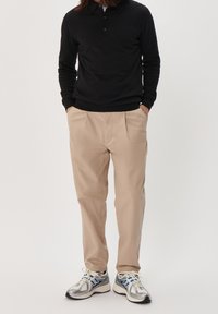 Polo noir à manches longues, pantalon beige avec poches latérales, et chaussures de sport argentées avec des accents bleus. Design simple et épuré.