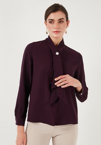 Blusa viola a maniche lunghe con colletto a cravatta, chiusura frontale con bottoni e sottili pieghe frontali. Tessuto liscio con una leggera lucentezza.
