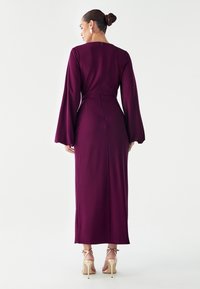 Strak, lang mouw jurk in diep bordeauxrode stof, voorzien van een ritssluiting aan de achterkant en een tot op de vloer reikend ontwerp met klokvormige mouwen.