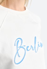 Bijela dukserica s lagano plavim izvezenim natpisom "Berlin". Dizajn uključuje okrugli izrez i mekanu teksturu tkanine.