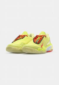 Een paar felgele sportschoenen met rode veters en gestructureerde beige zolen tegen een effen witte achtergrond.