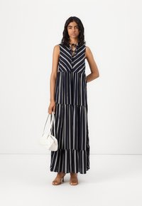 Vestido maxi listrado em azul-marinho com design em camadas, sem mangas e detalhe de laço no pescoço. Usado com sandálias de tiras em tom caramelizado e uma bolsa branca.