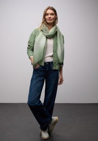 Veste bomber vert clair, haut blanc et jean large bleu foncé. La tenue comprend une écharpe verte texturée et des baskets blanches.