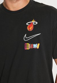 Camiseta negra de algodón que presenta el colorido logo de Miami Heat, el swoosh blanco de Nike y el texto "miami" en colores brillantes, todo de forma destacada en la parte frontal.