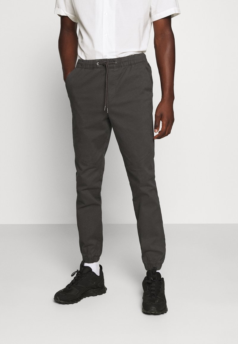 Mann trägt dunkelgraue Jogginghosen mit Kordelzug und elastischen Bündchen, weiße Socken, schwarze Turnschuhe und ein weißes kurzärmeliges Hemd mit Knöpfen.