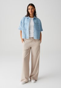 Chemise en denim bleu clair à manches courtes avec deux poches avant, associée à un pantalon large beige et des chaussures blanches. Une tenue simple et décontractée.