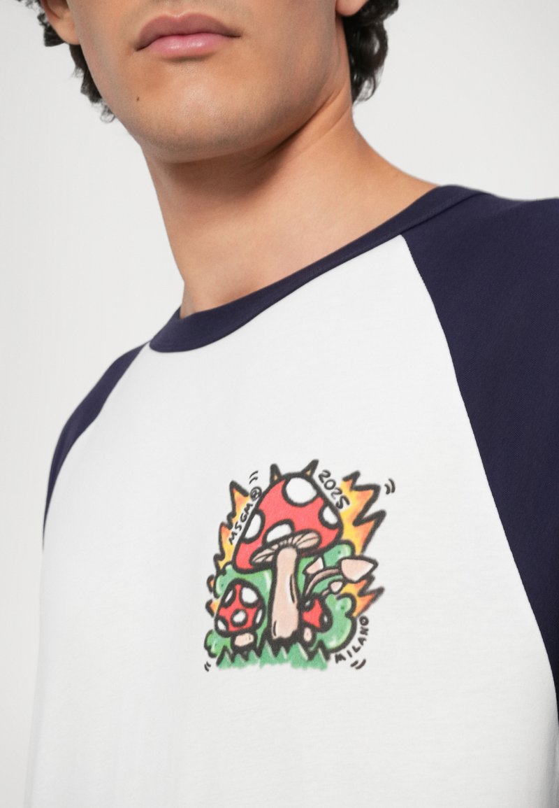 T-shirt blanc avec des manches bleu marine mettant en avant un graphique de champignon coloré, entouré de noir, avec deux champignons et des détails d'herbe verte.