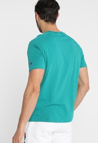 Muž otočený zády, oblečený v tyrkysovém tričku s krátkým rukávem a bílých šortkách s logy na rukávech a noze.