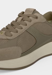 Baskets en mesh et daim beige et olive avec lacets assortis, étiquette dorée sur les lacets et semelle blanche épaisse.