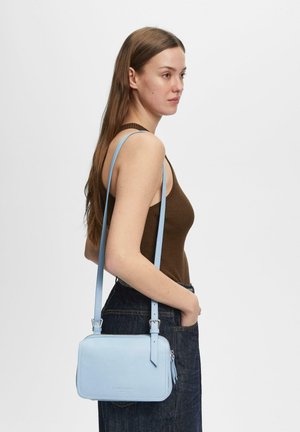 Kobieta w brązowym topie bez rękawów i ciemnych dżinsach, niosąca jasnobłękitną prostokątną torbę na ramię z regulowanym paskiem.