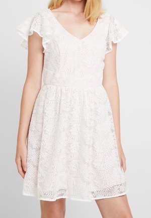 Vrouw draagt een witte kanten jurk met een V-hals, wijde mouwen en een geborduurd bloemenpatroon, staand tegen een effen achtergrond.