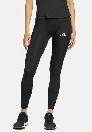 Vrouw draagt zwarte sportlegging met wit Adidas-logo op de dij en zwarte hardloopschoenen, staand tegen een witte achtergrond.