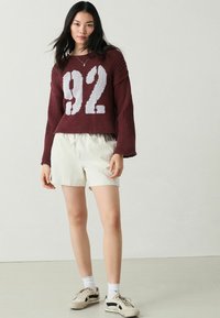 Vinröd stickad tröja med ett stort vitt tryck av "92", tillsammans med ljusa randiga shorts. Vita strumpor och beige sneakers fulländar outfiten.
